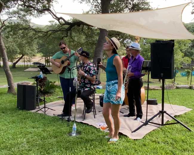 A band performs outdoors on a grassy area under a shaded canopy, with a singer in a swimsuit and three musicians playing various instruments.