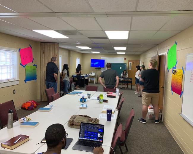 A group of people stands in a meeting room with a projector screen, while a long table is set with books and laptops.
