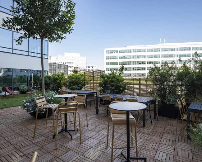 The image shows a rooftop terrace with several tables and chairs surrounded by greenery and buildings in the background.