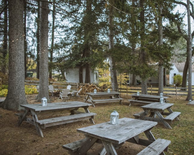 The image shows a wooded area with several wooden picnic tables surrounded by trees, leading to a white house in the background.
