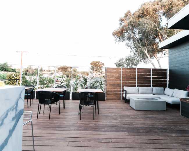 A wooden deck area furnished with tables and seating surrounded by greenery and modern fencing.