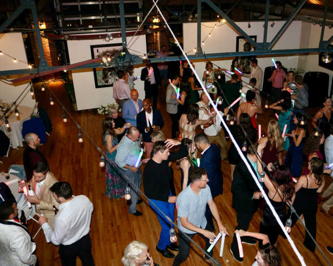 A large group of people dances with glow sticks under string lights in a spacious indoor venue.