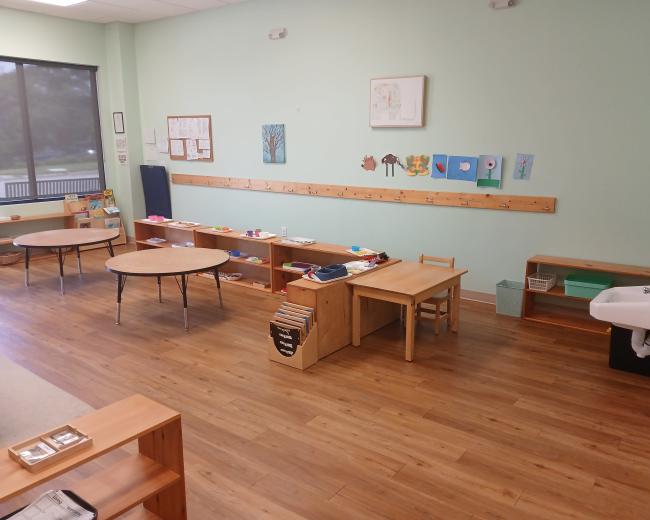 The image shows a classroom with wooden furniture, tables, shelves displaying various items, and artwork on the walls, set in a well-lit space with a window.