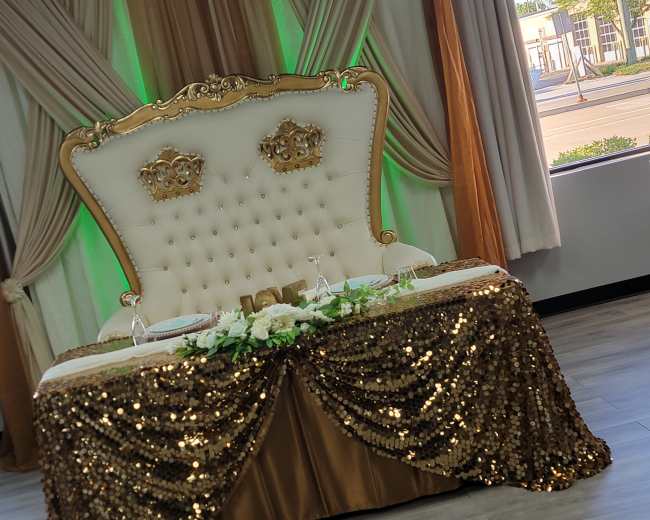 An elegantly decorated table with a gold sequined tablecloth, a tufted white and gold backdrop, and two crown accents on the chairs.