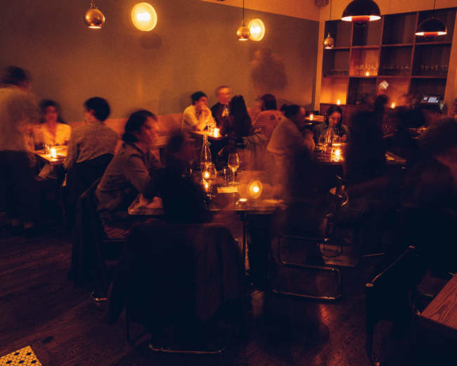The image shows a dimly lit restaurant filled with people dining at tables, creating a lively atmosphere.