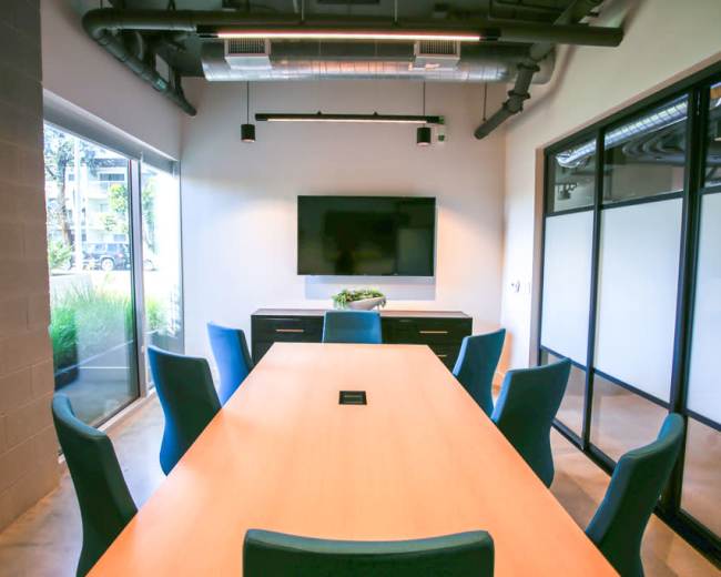 A conference room features a long wooden table surrounded by blue chairs, with a television mounted on the wall and large windows allowing natural light.