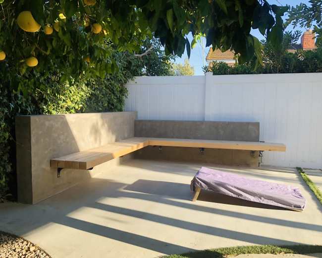 The image shows a concrete seating area enclosed by a white fence, with a wooden bench and a purple cushion on the ground under shaded greenery.