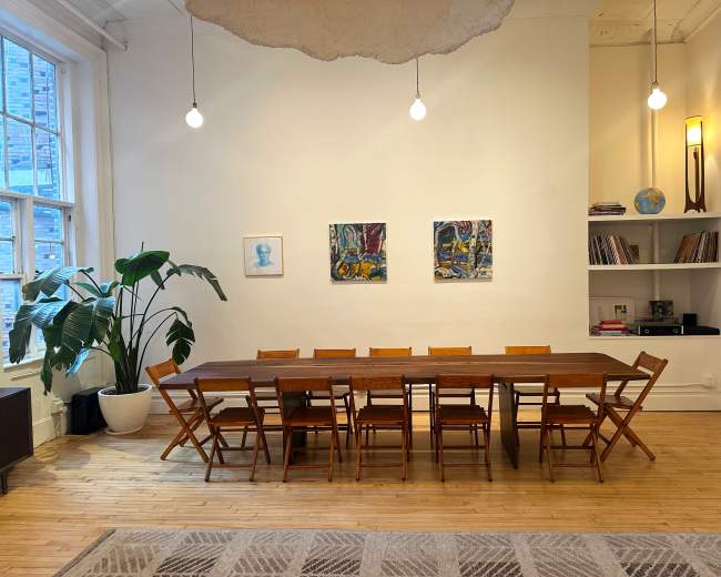 The image shows a spacious room with a large wooden dining table surrounded by folding chairs, two artworks on the wall, a tall plant in a pot, and shelves with books and a globe.