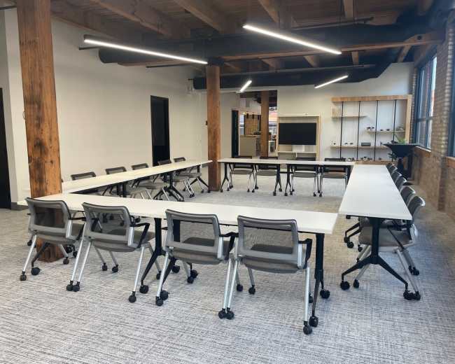 A modern conference room features a U-shaped arrangement of tables surrounded by gray rolling chairs, with exposed wooden beams and large windows.