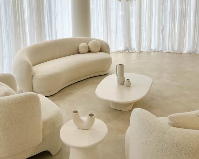 A modern, minimalist living space features white, curved furniture and sheer curtains, with a decorative light fixture overhead.