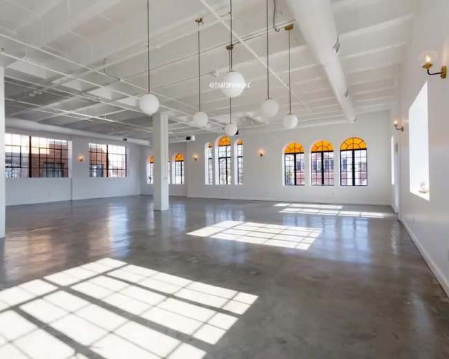 The image shows a spacious, empty room with large windows and bright, natural light casting shadows on the floor.