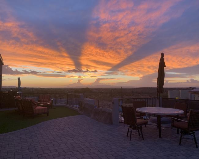 The scene shows a patio with furniture overlooking a vibrant sunset with long shadows stretching across the sky.