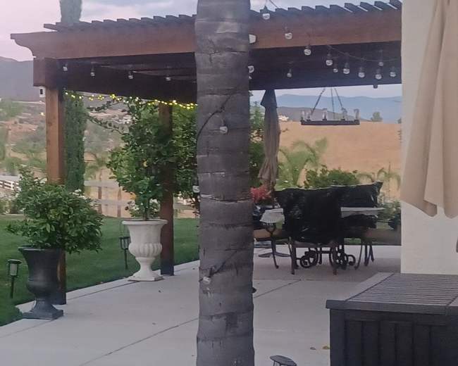 The image shows a landscaped backyard with potted plants, a patio area, and distant rolling hills.