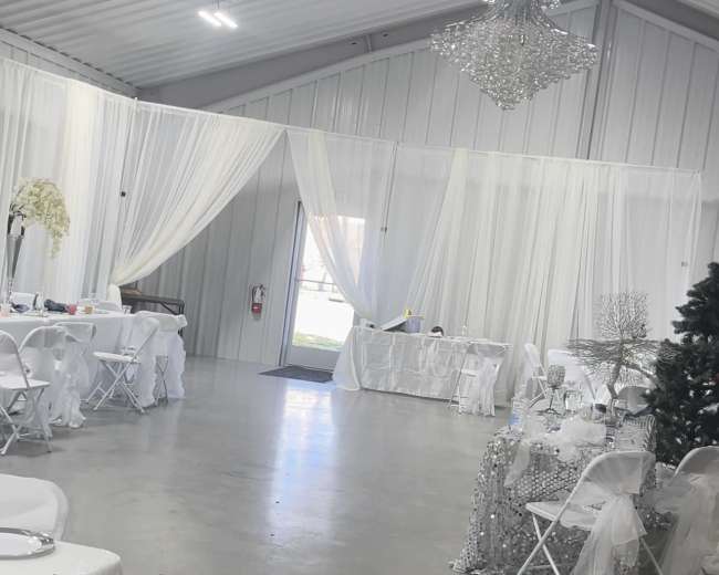 The image shows a spacious indoor venue decorated with white drapes and tables, featuring a chandelier and a Christmas tree.
