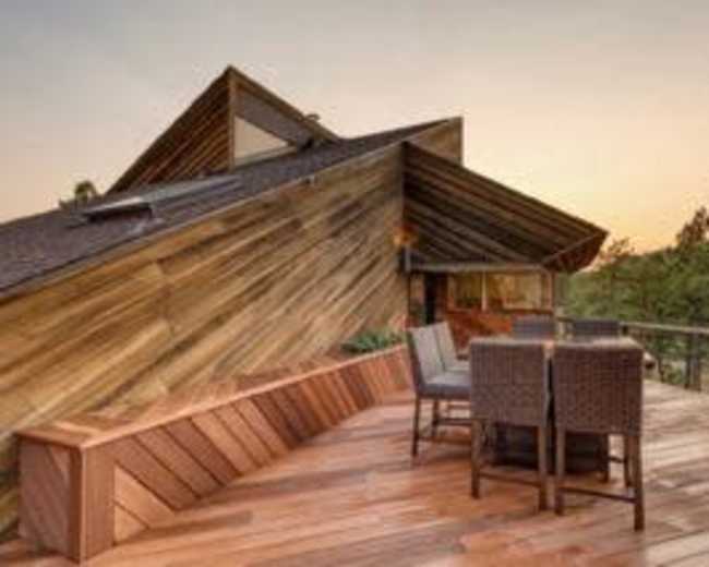 A wooden deck with a dining set is positioned next to a modern house featuring an angular roof design, surrounded by trees at sunset.