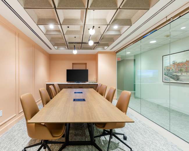 The image shows a modern conference room featuring a long wooden table with eight brown chairs, a large screen mounted on the wall, and glass panels providing a view into an adjoining space.