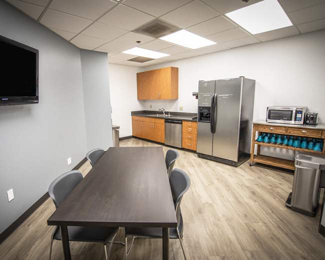 The image shows a break room with a table, chairs, a refrigerator, a microwave, and wooden cabinets against the wall.