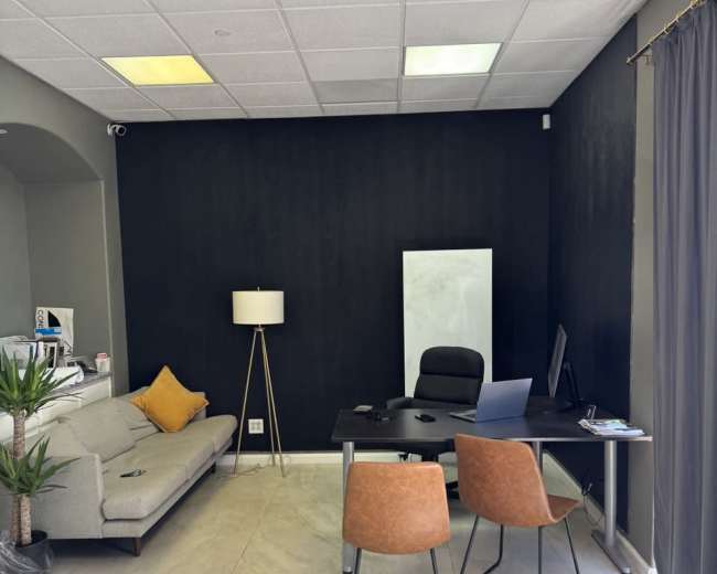The image shows a modern office space featuring a gray sofa, a floor lamp, a desk with a computer, and two orange chairs, all set against a black wall.
