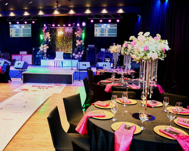 A decorated event space features tables set with black linens, pink accents, and elegant centerpieces, along with a stage adorned with lights and a backdrop.
