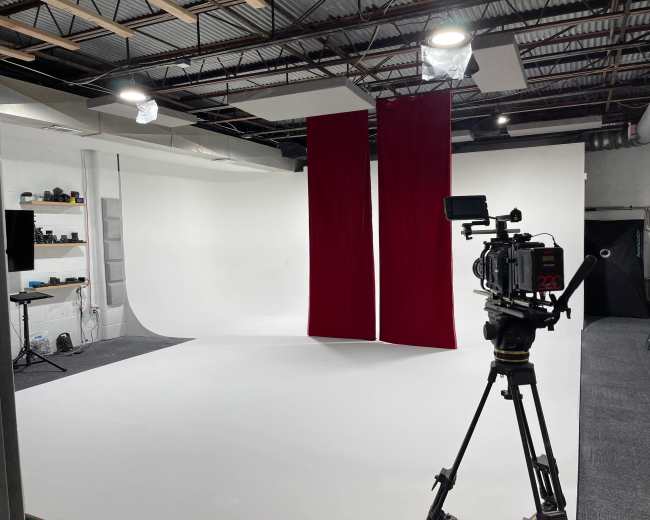 A photography studio with a white backdrop, two red curtains, and a camera on a tripod positioned in the foreground.