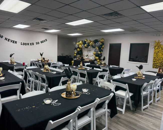 The image shows a decorated indoor event space with multiple tables set for a celebration, featuring black tablecloths, gold and silver decorations, and a backdrop for photos.