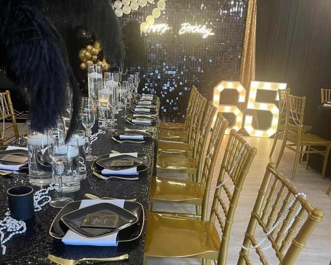 A lavish birthday banquet table is set with black and gold decorations, featuring elegant tableware, and illuminated numbers "65" in the background.