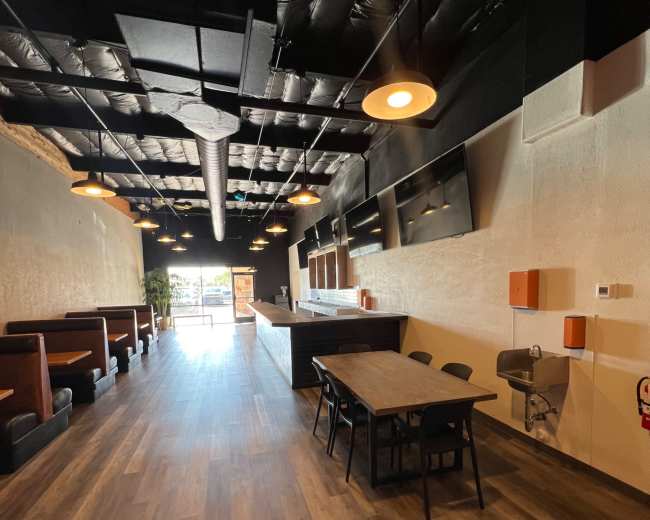 The image shows a spacious, modern restaurant interior with a long, narrow layout featuring wooden flooring, booths along the walls, and a central dining table.