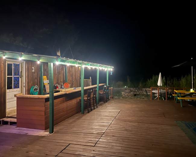 A wooden bar with stools is illuminated by string lights on a deck at night, surrounded by tall grass and a few seating options.