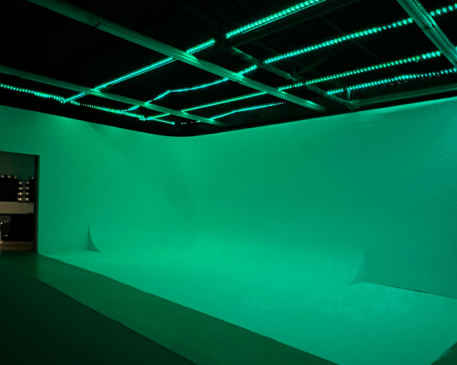 A studio space features a green backdrop and illuminated ceiling lights, creating a uniform color effect.
