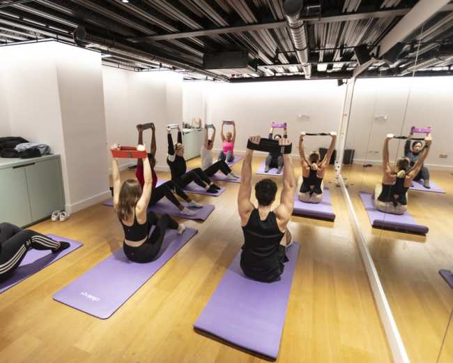 A group of individuals are participating in a fitness class, performing overhead exercises on yoga mats in a mirrored studio.