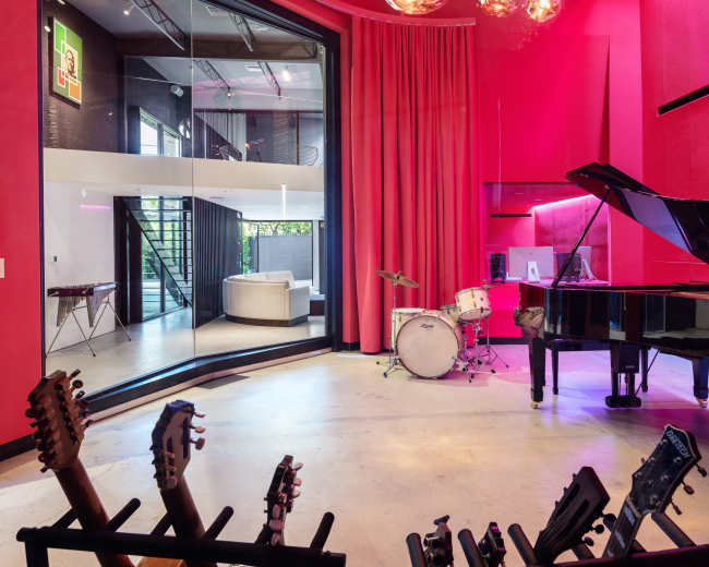 A vibrant music studio features a grand piano, drums, and several guitars displayed on stands, with large windows providing a view of an adjacent living space.