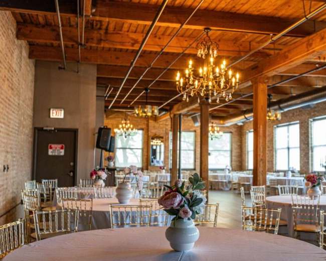 The image shows a spacious event venue with wooden beams, chandeliers, and round tables set with floral centerpieces.