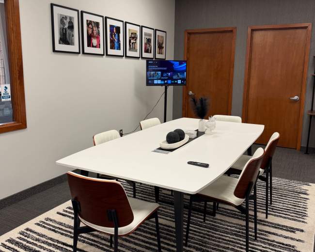 The image depicts a modern conference room with a large white table surrounded by six chairs, a television mounted on the wall, and framed photographs hanging above the table.
