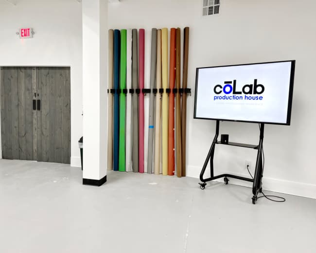 The image shows a spacious room with a large screen displaying the logo of "coLab production house" next to a colorful arrangement of backdrop rolls mounted on the wall.