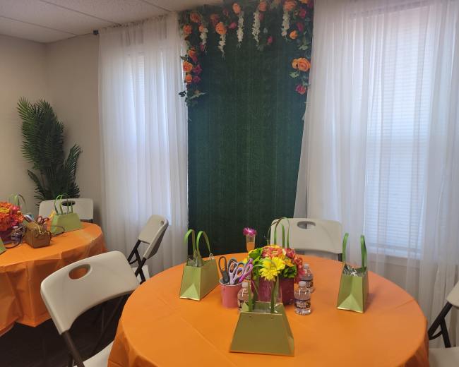 The image shows a decorated room with orange tablecloths, floral centerpieces, and green bags on round tables, against a backdrop of flowers and greenery.