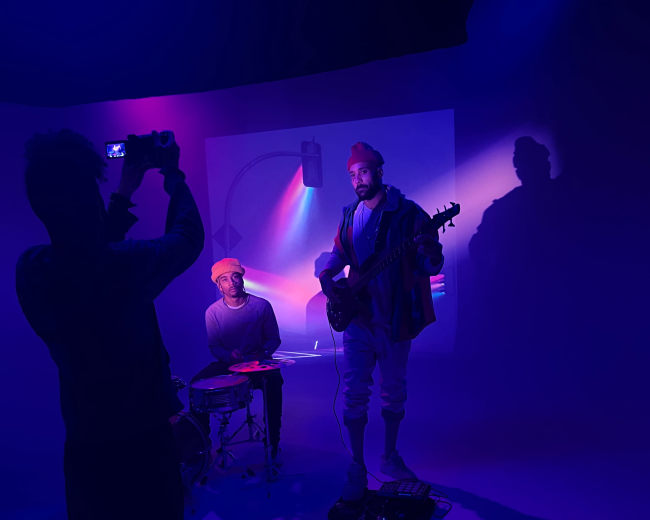 A musician plays guitar while another sits at a drum set, illuminated by colored lights in a studio setting.