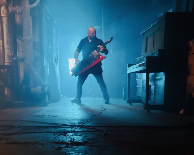 A musician plays a keytar surrounded by fog and dim lighting in an industrial setting.