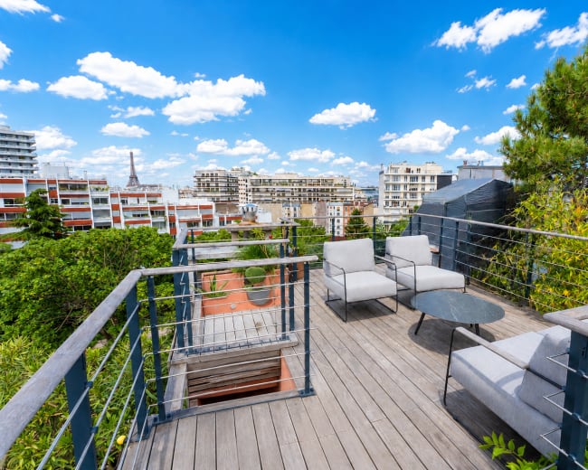 The image shows a rooftop terrace with modern furniture, including two chairs and a table, surrounded by greenery and city buildings under a blue sky with clouds.