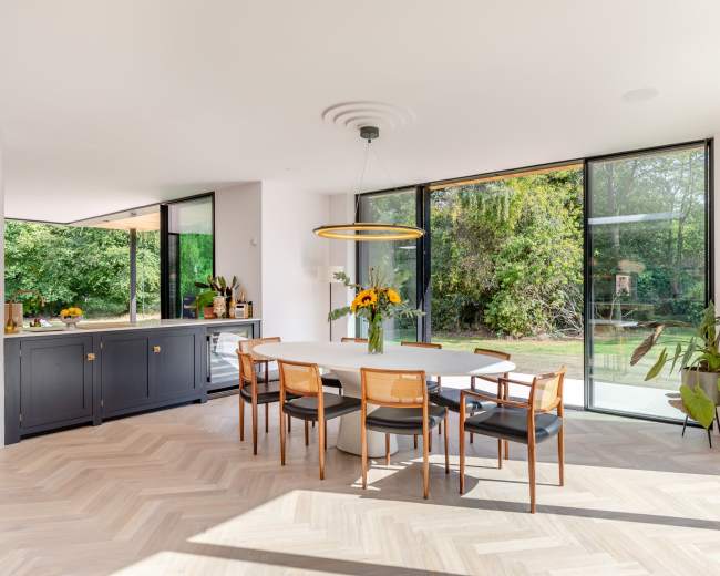 The image shows a modern dining area with a round white table surrounded by six chairs, large windows showcasing a garden view, and a side cabinet with decorative plants.