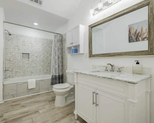 The image shows a bathroom featuring a bathtub with a tiled wall, a white vanity with a marble countertop, a mirror, and a toilet.