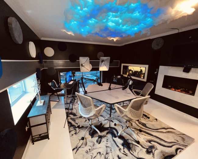 A modern studio space with a large table, chairs, and lighting equipment, featuring a textured rug and artistic ceiling design.