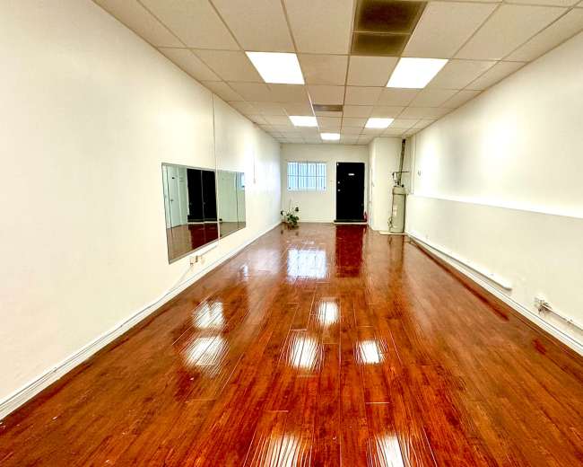 A spacious room with polished wooden floors, white walls, a large mirror on one side, and a door at the far end.