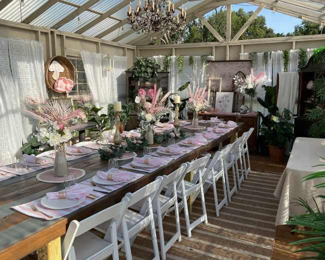 The image shows a decorated dining area with a long wooden table set for a meal, surrounded by plants and light-colored curtains in a greenhouse setting.