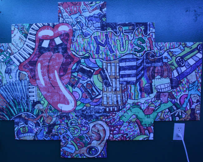 A large, colorful mural made up of various artistic motifs, including musical elements and abstract designs, mounted on a dark green wall.