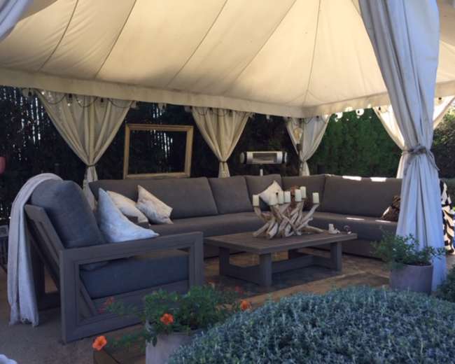 The image shows a shaded outdoor seating area with a large gray sectional sofa, a wooden coffee table, and decorative items, all under a tent-like structure.