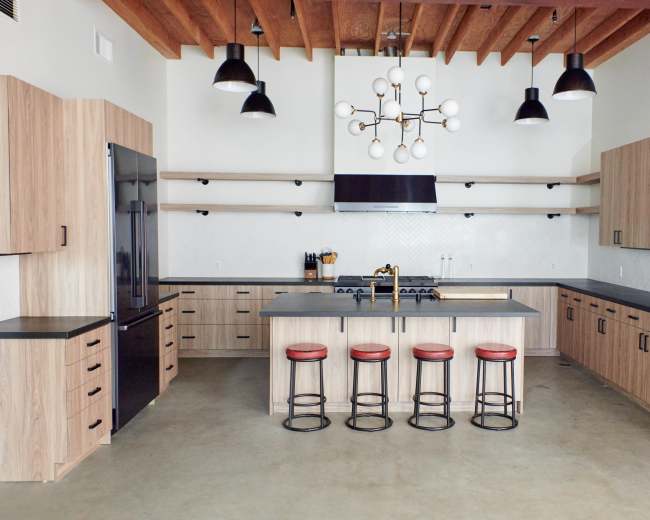 The image shows a modern kitchen with wooden cabinetry, black countertops, and four red-stool seating at an island.