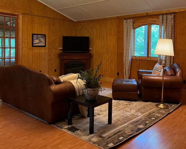 The image shows a cozy living room with brown leather sofas, a dark coffee table, a lamp, and wooden paneling on the walls.