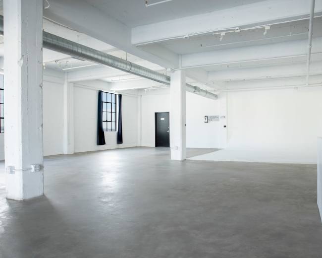 The image shows a spacious, empty interior featuring high ceilings, polished concrete floors, and large windows.