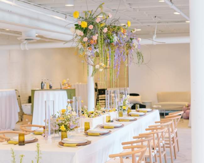 A long dining table is set with floral centerpieces and yellow accents in a spacious, well-lit room.