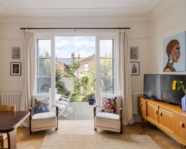 The image shows a bright living room with large windows overlooking a garden, featuring a dining table, two armchairs, a television, and various wall art.